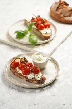 Kızarmış kiraz domatesli sandviç, taze süzme peynir, yeşil fesleğen, gri arka planda yuvarlak bir tabakta kepekli ekmek dilimi.