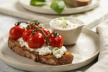 Kızarmış kiraz domatesli sandviç, taze süzme peynir, yeşil fesleğen, gri arka planda yuvarlak bir tabakta kepekli ekmek dilimi.