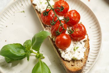 Kızarmış kiraz domatesli sandviç, taze süzme peynir, yeşil fesleğen, gri arka planda yuvarlak bir tabakta kepekli ekmek dilimi.