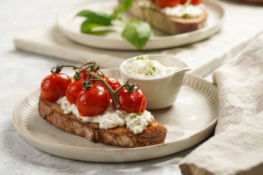 Kızarmış kiraz domatesli sandviç, taze süzme peynir, yeşil fesleğen, gri arka planda yuvarlak bir tabakta kepekli ekmek dilimi.