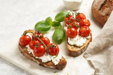 A sandwich with roasted cherry tomatoes with branch, fresh cottage cheese, green basil on a slice of whole wheat bread on a marble tray on grey background