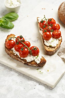 A sandwich with roasted cherry tomatoes with branch, fresh cottage cheese, green basil on a slice of whole wheat bread on a marble tray on grey background