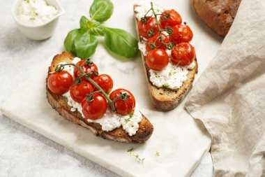 Kızarmış kiraz domatesli sandviç, taze süzme peynir, yeşil fesleğen, gri arka planda mermer tepside kepekli tam buğday ekmeği.