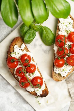 Kızarmış kiraz domatesli sandviç, taze süzme peynir, yeşil fesleğen, yeşil bitki dallarıyla çevrili yeşil arkaplanda mermer tepside kepekli tam buğday ekmeği.