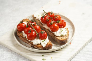 Kızarmış kiraz domatesli sandviç, taze süzme peynir, kepekli yeşil fesleğen, gri arkaplanda mermer tepside yuvarlak bir tabakta kepekli ekmek.