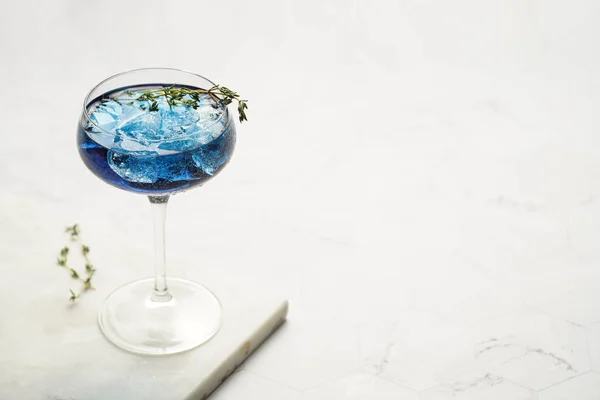 Dark blue drink in a vintage glass for sparkling wine - pea flower tea or blue curacao sirup cocktail with thyme branches on marble board. Copy space for text