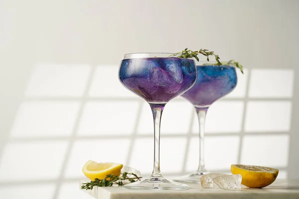 a blue drink in a vintage glass for sparkling wine slowly becoming purple - pea flower tea or blue curacao sirup cocktail with thyme branches on white background. Copy space for text