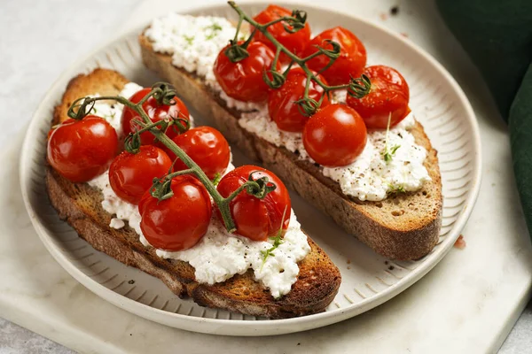 a sandwich with roasted cherry tomatoes with branch, fresh cottage cheese, green basil on a slice of whole wheat bread on a round plate on marble tray on grey background