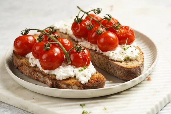 a sandwich with roasted cherry tomatoes with branch, fresh cottage cheese, green basil on a slice of whole wheat bread on a round plate on marble tray on grey background