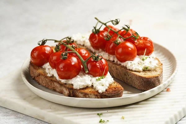 a sandwich with roasted cherry tomatoes with branch, fresh cottage cheese, green basil on a slice of whole wheat bread on a round plate on marble tray on grey background