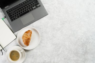 A croissant and a cup of coffee on white plates on grey working desk in the office with laptop and notebook, copy space for text