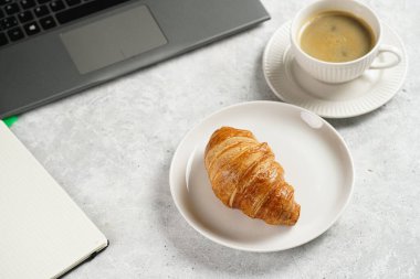 A croissant and a cup of coffee on white plates on grey working desk in the office with laptop and notebook, copy space for text