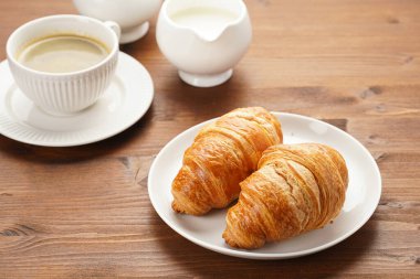 Two french soft croissants on a white plate on a wooden table, a cup of black coffee in white sophisticated cup, milk, sugar, jam