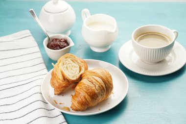 Two french soft croissants on a white plate on a blue wooden table, a cup of black coffee in white sophisticated cup, milk, sugar, jam