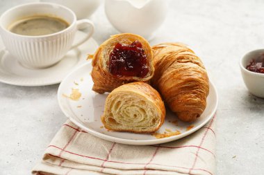 Two french soft croissants with jam on a white plate on a checkered napkin, a cup of black coffee in white sophisticated cup, milk, sugar