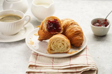 Two french soft croissants with jam on a white plate on a checkered napkin, a cup of black coffee in white sophisticated cup, milk, sugar