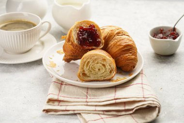 Two french soft croissants with jam on a white plate on a checkered napkin, a cup of black coffee in white sophisticated cup, milk, sugar