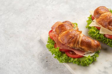 two lye croissant sandwiches with iberian ham, tomato slices, lettuce and cheese on marble board on grey background