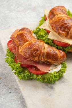 two lye croissant sandwiches with iberian ham, tomato slices, lettuce and cheese on marble board on grey background