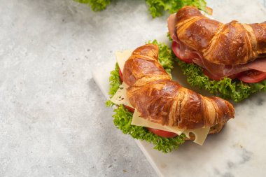 two lye croissant sandwiches with iberian ham, tomato slices, lettuce and cheese on marble board on grey background