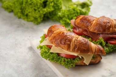 two lye croissant sandwiches with iberian ham, tomato slices, lettuce and cheese on marble board on grey background