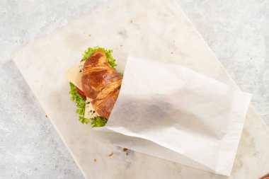 lye croissant sandwich with iberian ham, tomato slices, lettuce and cheese in a breakfast white paper bag on marble board on grey background