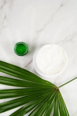 A mock-up of a round cosmetics container with cream on marble background and green palm leaf, top view