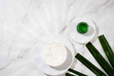 A mock-up of a round cosmetics container with cream on marble background and green palm leaf, top view