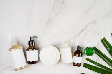 A mock-up of a brown cosmetics bottle with dispenser and white label, round cream container, bottle with pipette on marble background lined up and green palm leaf, top view