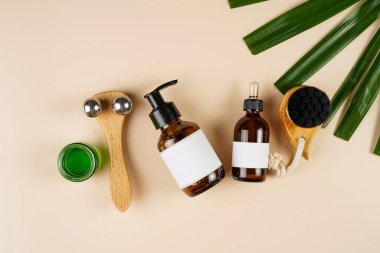A mock-up of a brown cosmetics bottle with dispenser and white label, round green cream container, bottle with pipette, face brush, face massage roller on beige colored background lined up, top view