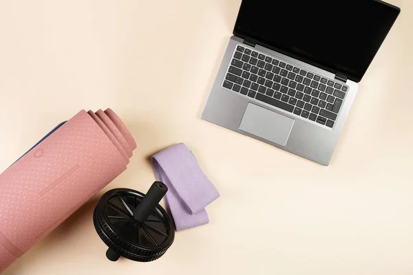 Clothes and equipment for a home workout or gym: pink yoga matt, purple fitness band, black abs-wheel, open laptop on beige colored background, top view, copy space