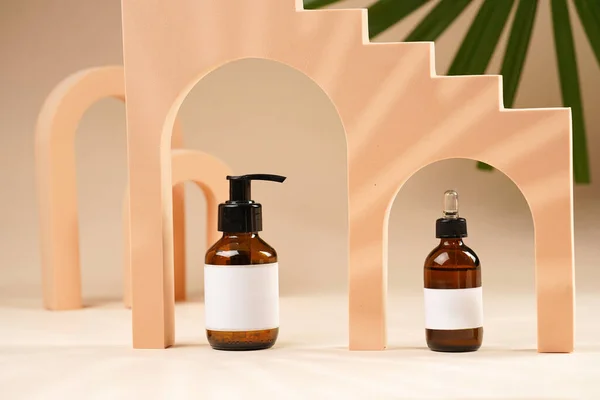 A mock-up of a brown cosmetics bottle with dispenser and white label and bottle with pipette in peach colored arched doors for cosmetics photography on beige colored background