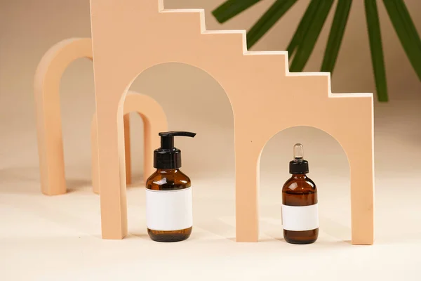 A mock-up of a brown cosmetics bottle with dispenser and white label and bottle with pipette in peach colored arched doors for cosmetics photography on beige colored background