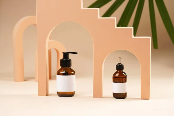 A mock-up of a brown cosmetics bottle with dispenser and white label and bottle with pipette in peach colored arched doors for cosmetics photography on beige colored background