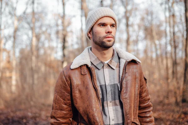 A portrait of a young white man posing in the woods dressed in autumn clothes: brown jacket, checkered canadian style shirt, off-white beanie, looking to the right