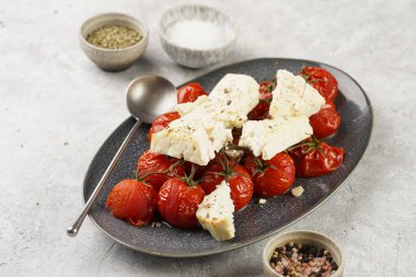 Roasted red tomatoes on branches with seasoned feta cheese on oval blue plate on grey background, top view