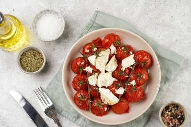 Roasted red tomatoes on branches with seasoned feta cheese on round off-white plate on kitchen napkins on grey background