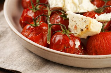 Roasted red tomatoes on branches with seasoned feta cheese on round off-white plate on kitchen napkins on wooden background
