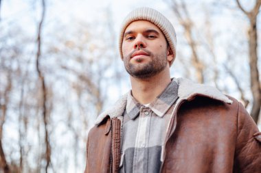 A portrait of a young white man posing in the woods dressed in autumn clothes: brown jacket, checkered canadian style shirt, off-white beanie, looking at the lens