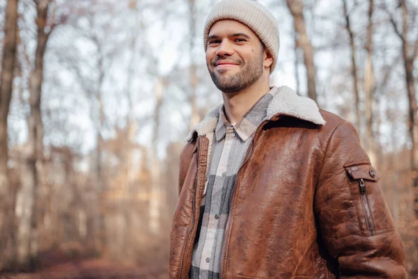 A portrait of a young white man posing in the woods dressed in autumn clothes: brown jacket, checkered canadian style shirt, off-white beanie, looking at the lens and smiling