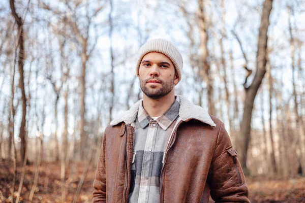 A portrait of a young white man posing in the woods dressed in autumn clothes: brown jacket, checkered canadian style shirt, off-white beanie, looking at the lens