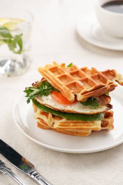 Savory breakfast - fresh homemade belgian waffle sandwich with green lettuce, crisp bacon and fried egg on white plate on beige colored table cloth, glass with lemon water, cup of coffee