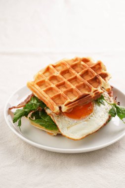 Savory breakfast - fresh homemade belgian waffle sandwich with green lettuce, crisp bacon and fried egg on white plate on beige colored table cloth
