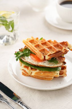 Savory breakfast - fresh homemade belgian waffle sandwich with green lettuce, crisp bacon and fried egg on white plate on beige colored table cloth, glass with lemon water, cup of coffee