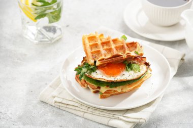 Savory breakfast - fresh homemade belgian waffle sandwich with green lettuce, crisp bacon and fried egg on white plate on beige colored table cloth, glass with lemon water, cup of coffee