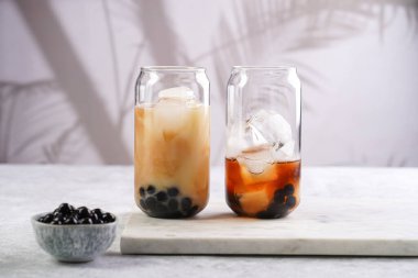 Two glasses with black tea, milk, ice cubes and cooked tapioca pearls for trendy bubble boba ice tea, two small grey ceramic bowls on marble board on pastel tropical background