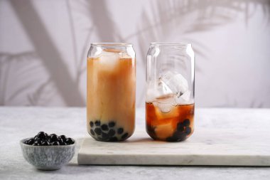 Two glasses with black tea, milk, ice cubes and cooked tapioca pearls for trendy bubble boba ice tea, two small grey ceramic bowls on marble board on pastel tropical background clipart