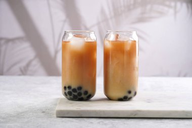 Two glasses with black tea, milk, ice cubes and cooked tapioca pearls for trendy bubble boba ice tea, two small grey ceramic bowls on marble board on pastel tropical background
