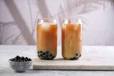 Two glasses with black tea, milk, ice cubes and cooked tapioca pearls for trendy bubble boba ice tea, two small grey ceramic bowls on marble board on pastel tropical background