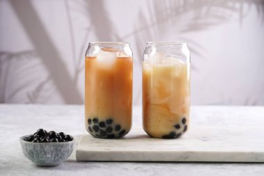 Two glasses with black tea, milk, ice cubes and cooked tapioca pearls for trendy bubble boba ice tea, two small grey ceramic bowls on marble board on pastel tropical background